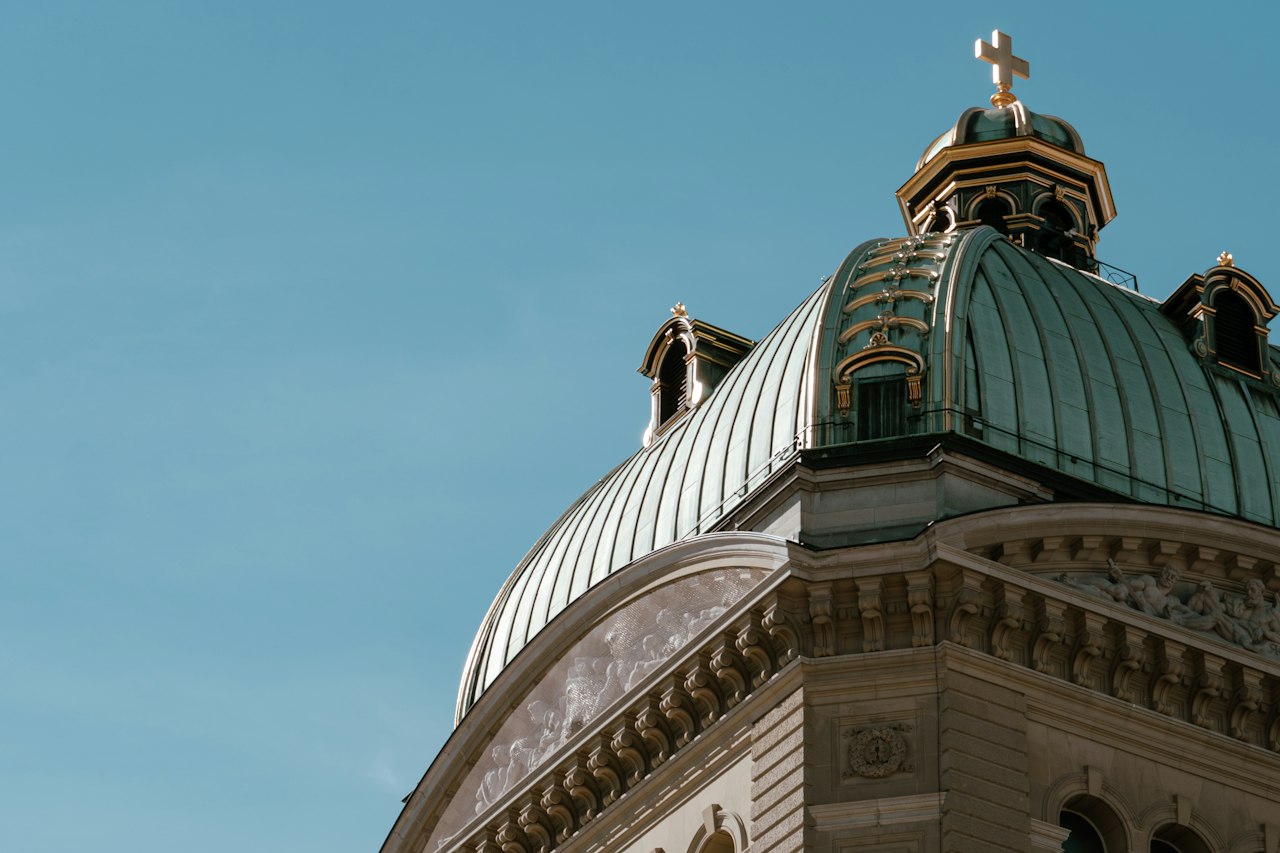 The ribbed copper dome of the Federal Palace in Bern, topped by a gilded cross, above a carved stone cornice against a clear sky